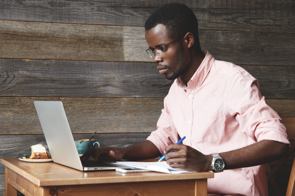 Successful African American businessman sitting at a cafe table, working on generic laptop, typing a message with one hand and filling in papers with a pen with the other hand. Business and technology