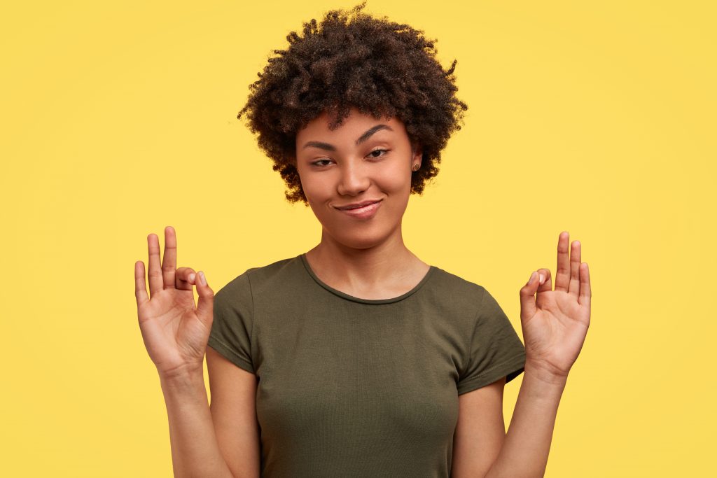 Delighted African American female shows ok sign, calms you that everything is fine, asks not worry, has positive confident expression, dressed in casual clothing, poses against yellow background